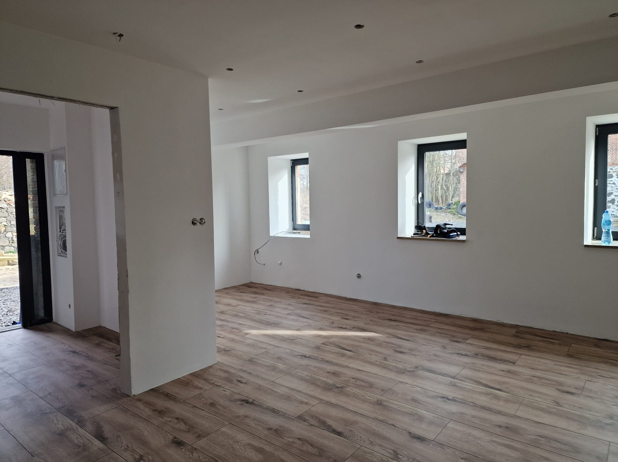 Wnętrze pokoju po remoncie z nową podłogą z paneli imitujących drewno, świeżo pomalowanymi ścianami na biało i zamontowanymi oknami z czarnymi ramami. Widoczne przygotowane otwory pod oświetlenie...