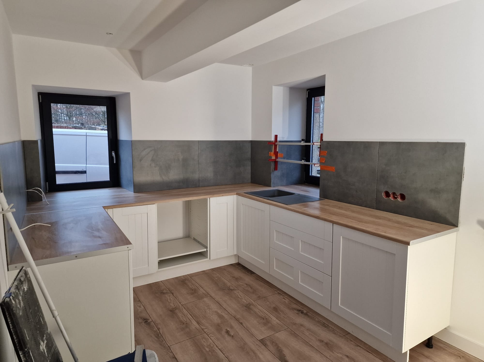 Kątowe ujęcie kuchni w trakcie prac: częściowo położone szare płytki nad blatem z imitacją drewna, białe szafki z frezowanymi frontami, widoczne otwory na gniazdka elektryczne i okno z czarną ramą.