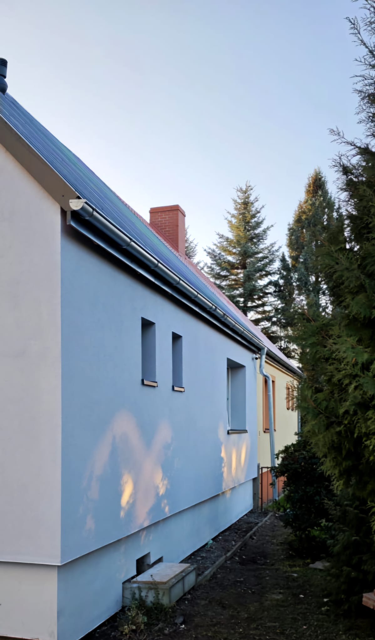 Świeżo pomalowana elewacja domu w odcieniach szarości, z widocznymi panelami słonecznymi na dachu, małymi oknami i refleksami słońca na ścianie, otoczona zielenią.