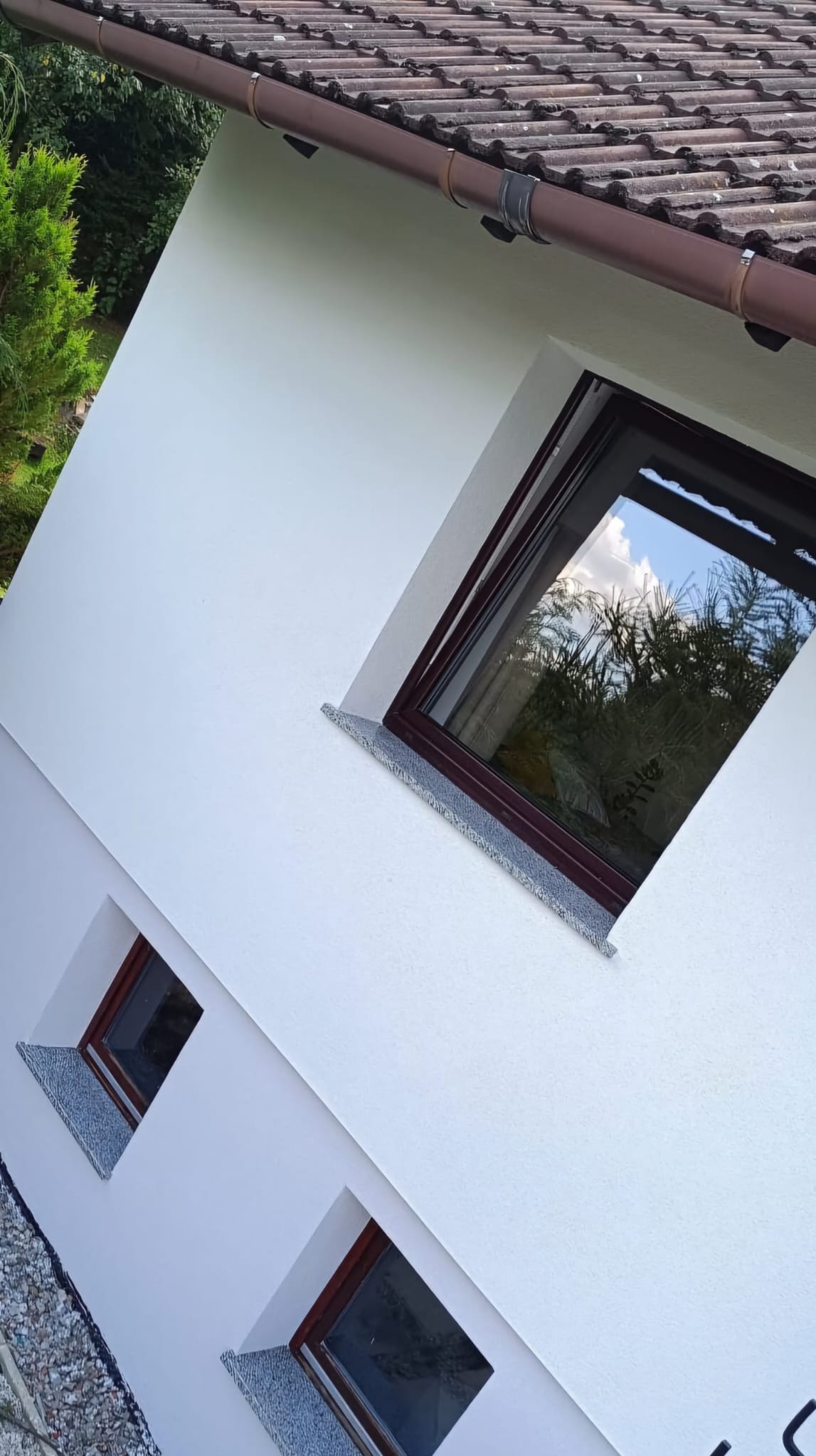 Świeżo pomalowana, biała elewacja domu z brązowymi oknami i parapetami wykończonymi szarym kamieniem, widoczne odbicie drzew w szybach okiennych i fragment brązowej rynny.