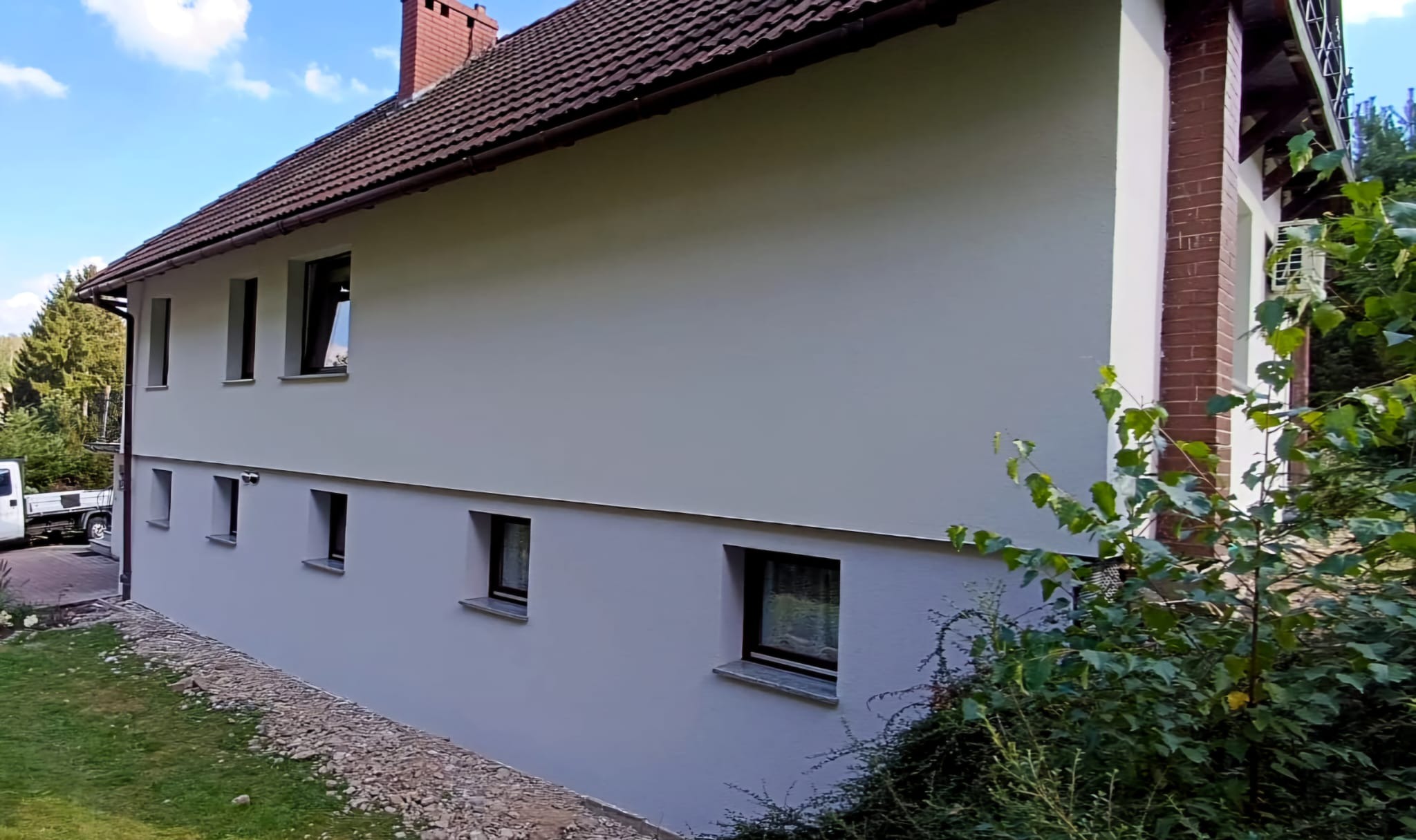 Elewacja domu po malowaniu na szaro, widoczne okna z ciemnymi ramami, dach z brązowej dachówki i fragment ogrodu z zielenią.