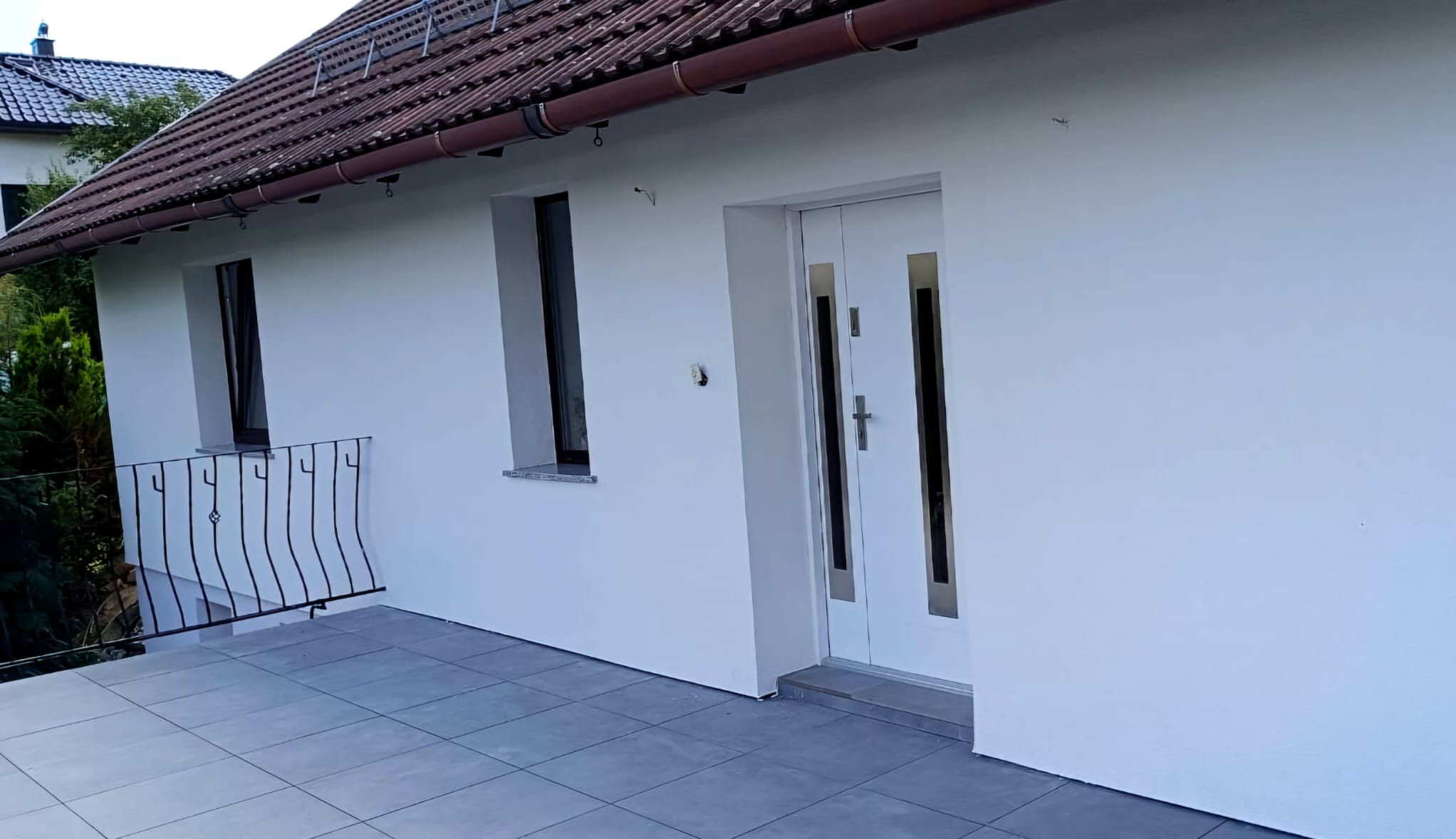 Biała elewacja domu z nowymi oknami i drzwiami wejściowymi, widoczny balkon z metalową balustradą i szara płytka na podłodze, brązowy dach z rynnami.