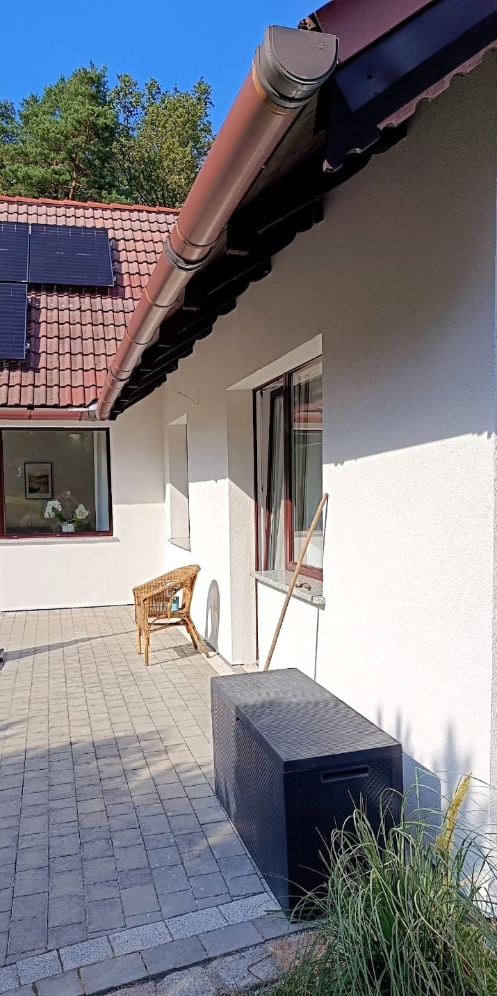 Odnowiona elewacja domu z brązowymi rynnami, panelami słonecznymi na dachu i wybrukowanym tarasem z wiklinowym krzesłem i skrzynią ogrodową