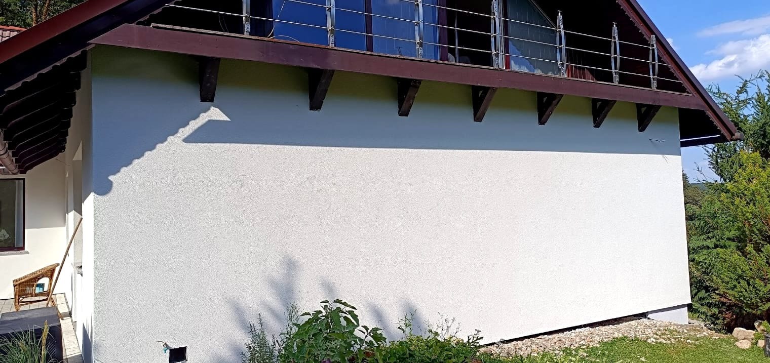 Boczna ściana domu po świeżym pomalowaniu na biało, widoczne brązowe elementy konstrukcyjne dachu, balkon z metalową barierką oraz zieleń w tle.