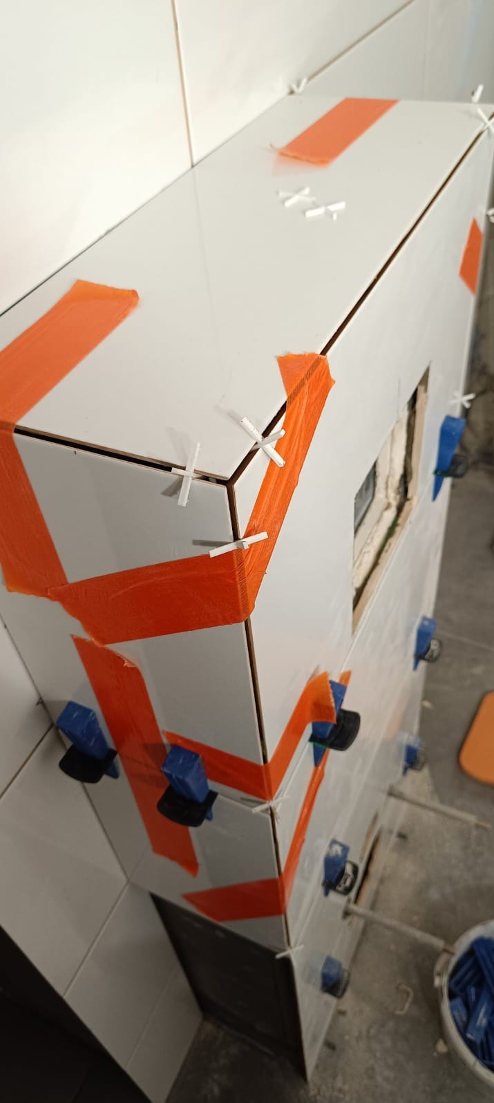 Białe płytki ceramiczne na obudowie w trakcie montażu, zabezpieczone pomarańczową taśmą i niebieskimi klipsami poziomującymi, z widocznymi krzyżykami dystansowymi i fragmentem ściany z boku.