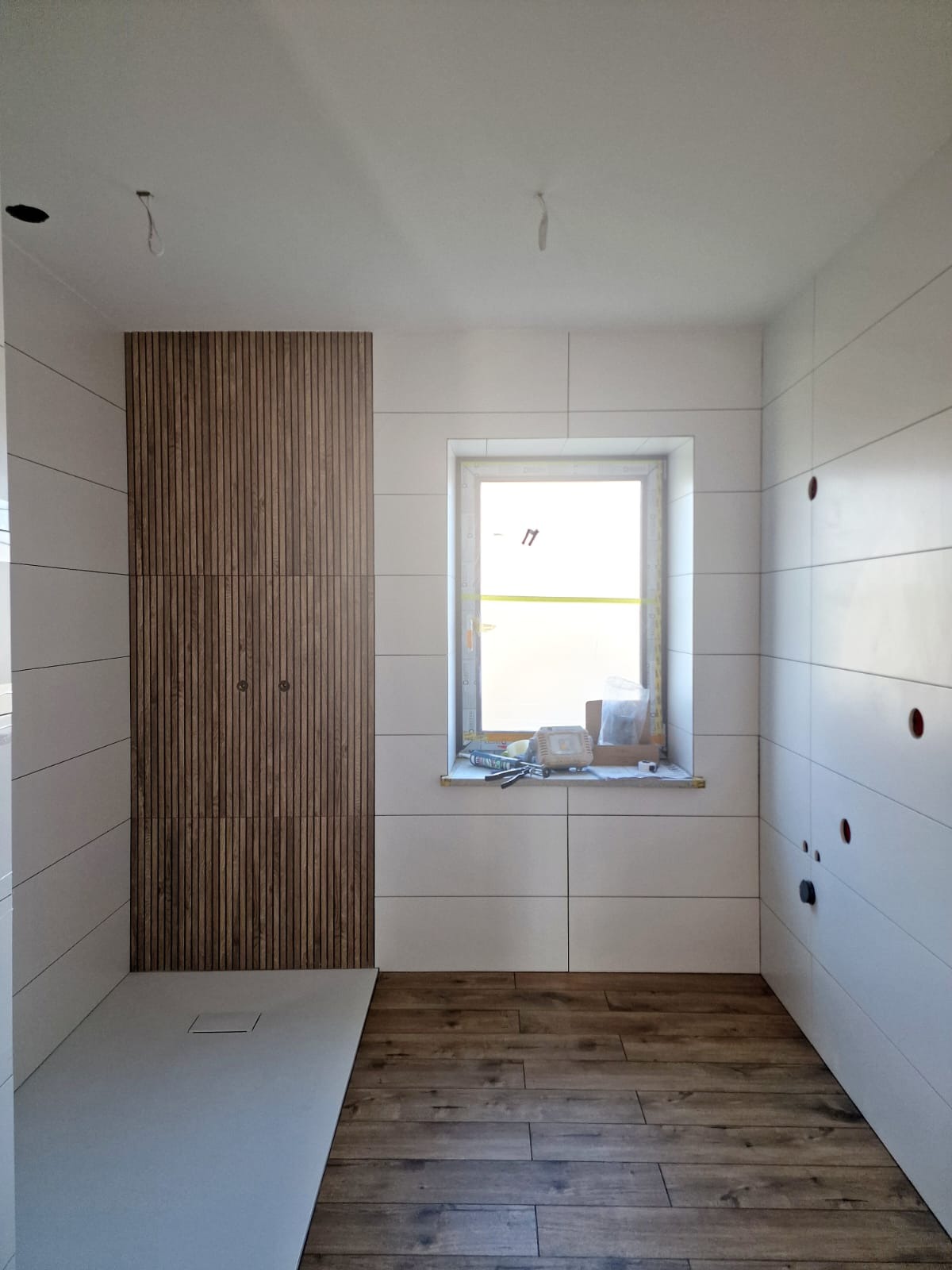 Wnętrze łazienki w trakcie wykańczania z widocznym brodzikiem, drewnianą ścianą dekoracyjną, białymi płytkami na ścianach, drewnopodobną podłogą oraz oknem z narzędziami na parapecie.