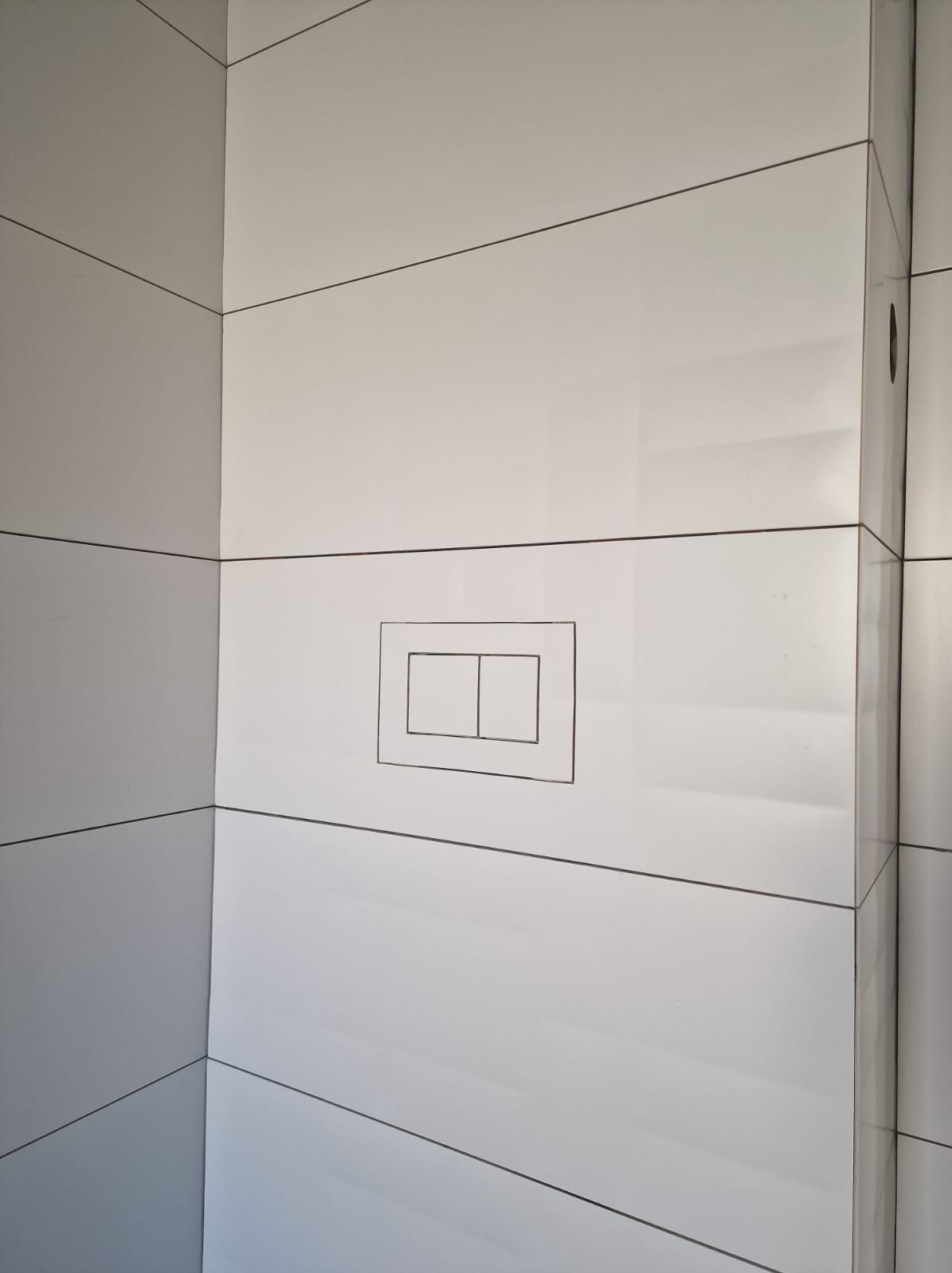 Białe, błyszczące płytki ceramiczne ułożone poziomo w narożniku łazienki, z widocznym przyciskiem spłuczki w zabudowie podtynkowej.