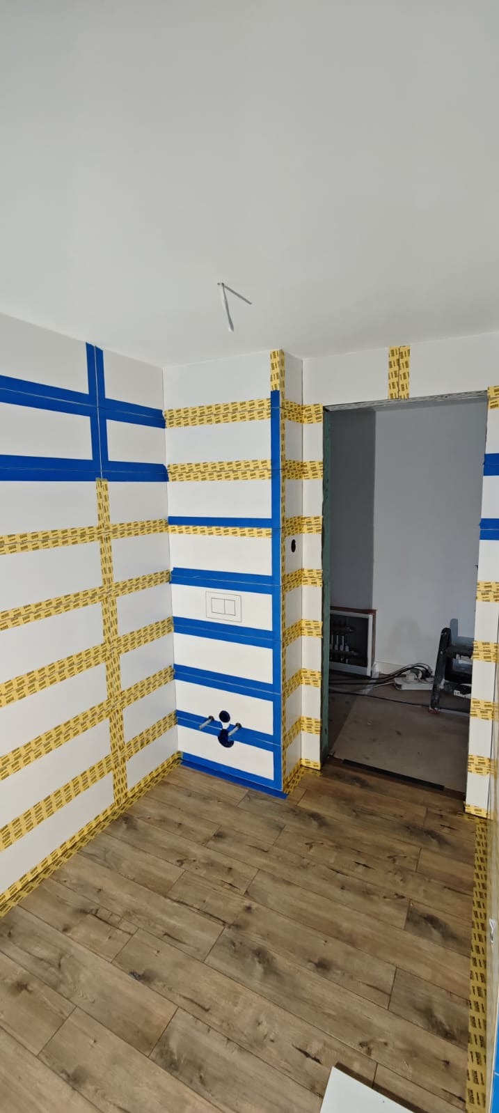 Łazienka w Zgorzelcu w trakcie prac wykończeniowych, ściany i obudowa stelaża WC zabezpieczone niebieską taśmą malarską i taśmą maskującą, widoczne przyłącza wodne i otwór na przycisk spłuczki,...