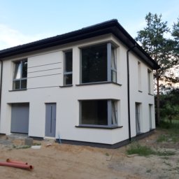 BARTOR - Dwukondygnacyjny dom z jasną elewacją, szarą stolarką okienną i ciemnym dachem, widoczny z zewnątrz na tle drzew i nieba, z elementami przygotowania terenu przed budynkiem.