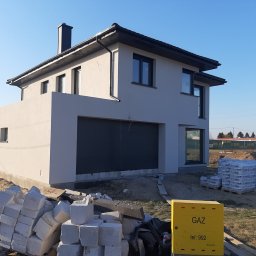 BARTOR - Nowoczesny dom jednorodzinny z szarą elewacją i grafitową bramą garażową, widoczne materiały budowlane i skrzynka gazowa.