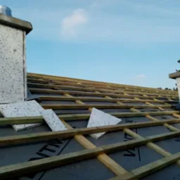 Poddasze w trakcie ocieplania: widoczne drewniane krokwie, czarna membrana, fragmenty białego styropianu oraz kominy z szarego betonu z metalowymi wkładami na tle błękitnego nieba.