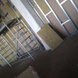 BARTOR - Pracownik na rusztowaniu montuje izolację z wełny mineralnej w metalowym stelażu na ścianie, widoczne płyty kartonowo-gipsowe.