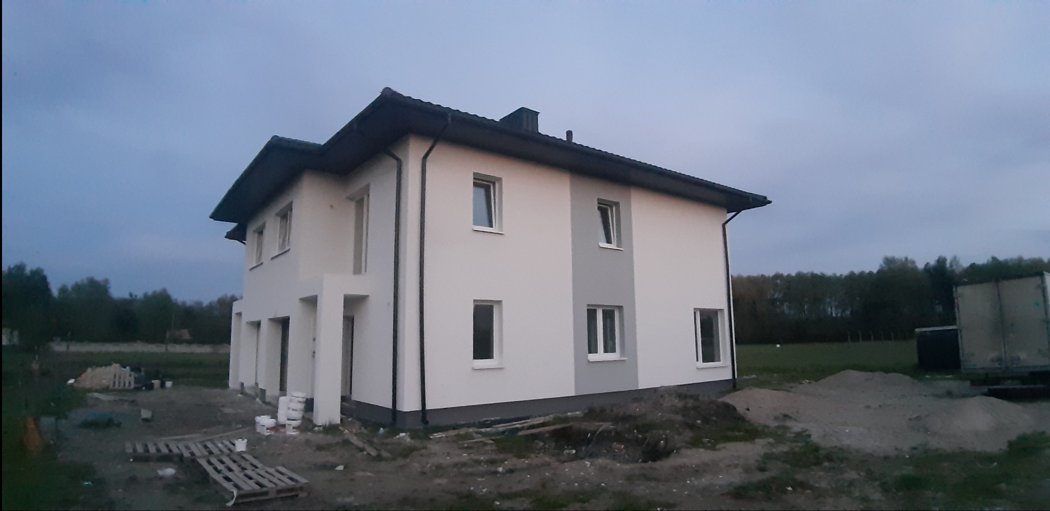 Dwukondygnacyjny dom jednorodzinny z jasną elewacją i szarym pasem, widoczny z boku, z elementami wykończeniowymi w kolorze czarnym, na działce z pozostałościami materiałów budowlanych.