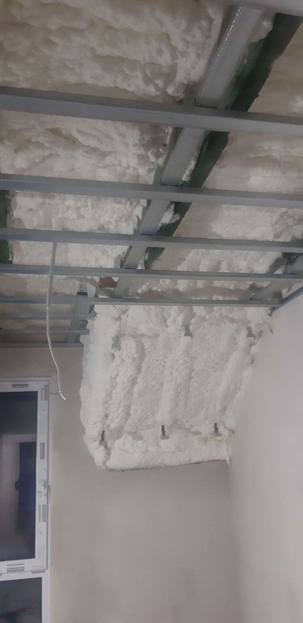 Poddasze z widoczną konstrukcją metalową i nałożoną białą pianą izolacyjną, częściowo zakrywająca ścianę, widoczne mocowania konstrukcji.