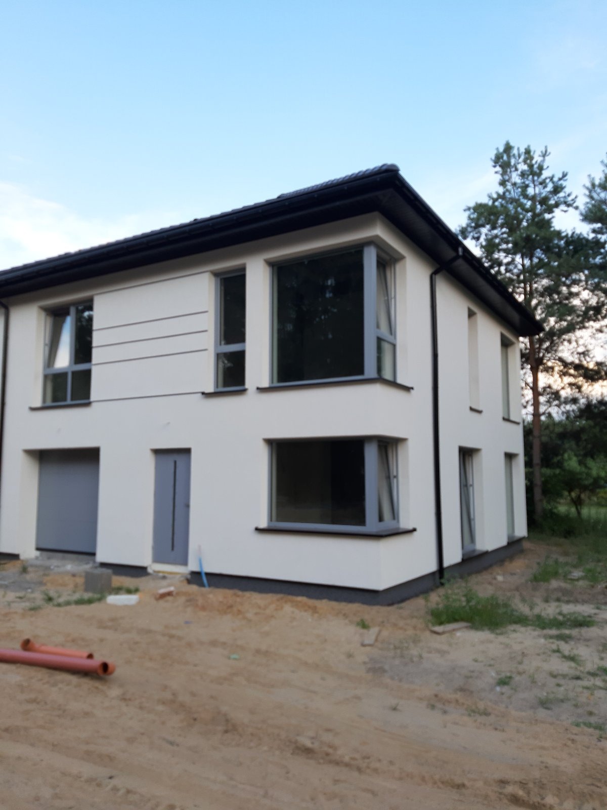 Dwukondygnacyjny dom z jasną elewacją, szarą stolarką okienną i ciemnym dachem, widoczny z zewnątrz na tle drzew i nieba, z elementami przygotowania terenu przed budynkiem.