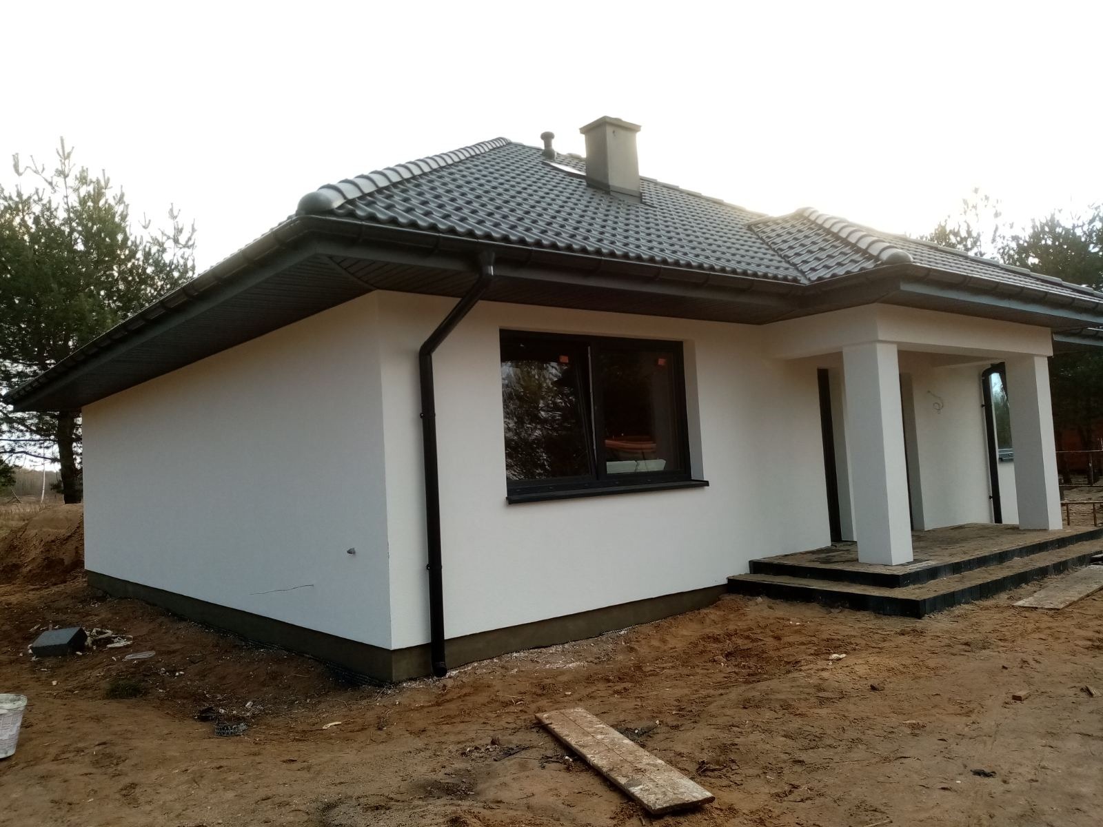 Nowo otynkowany dom jednorodzinny w Wieliszewie z grafitową dachówką i stolarką okienną, widoczne elementy wykończeniowe elewacji i zagospodarowania terenu.