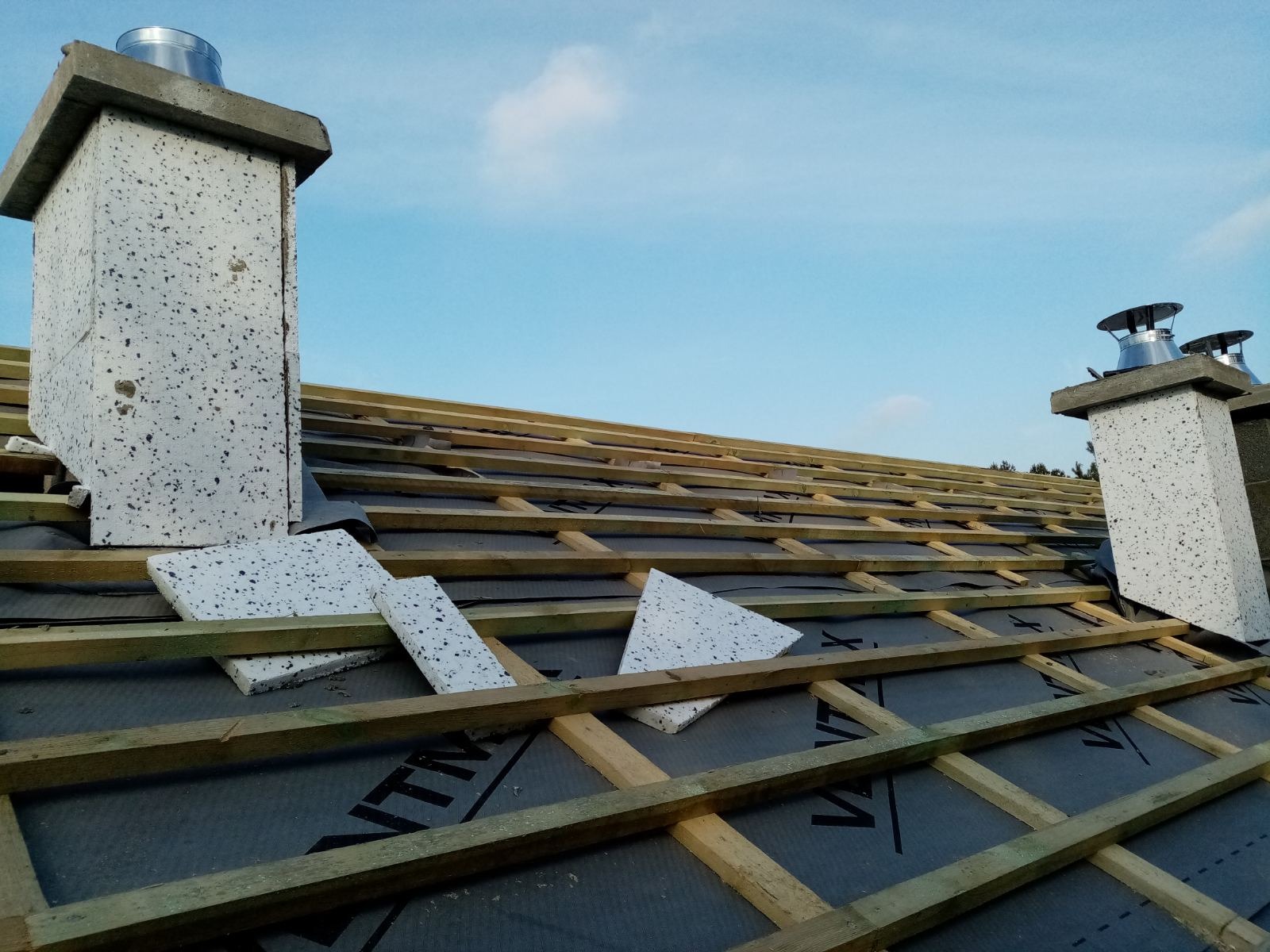 Poddasze w trakcie ocieplania: widoczne drewniane krokwie, czarna membrana, fragmenty białego styropianu oraz kominy z szarego betonu z metalowymi wkładami na tle błękitnego nieba.
