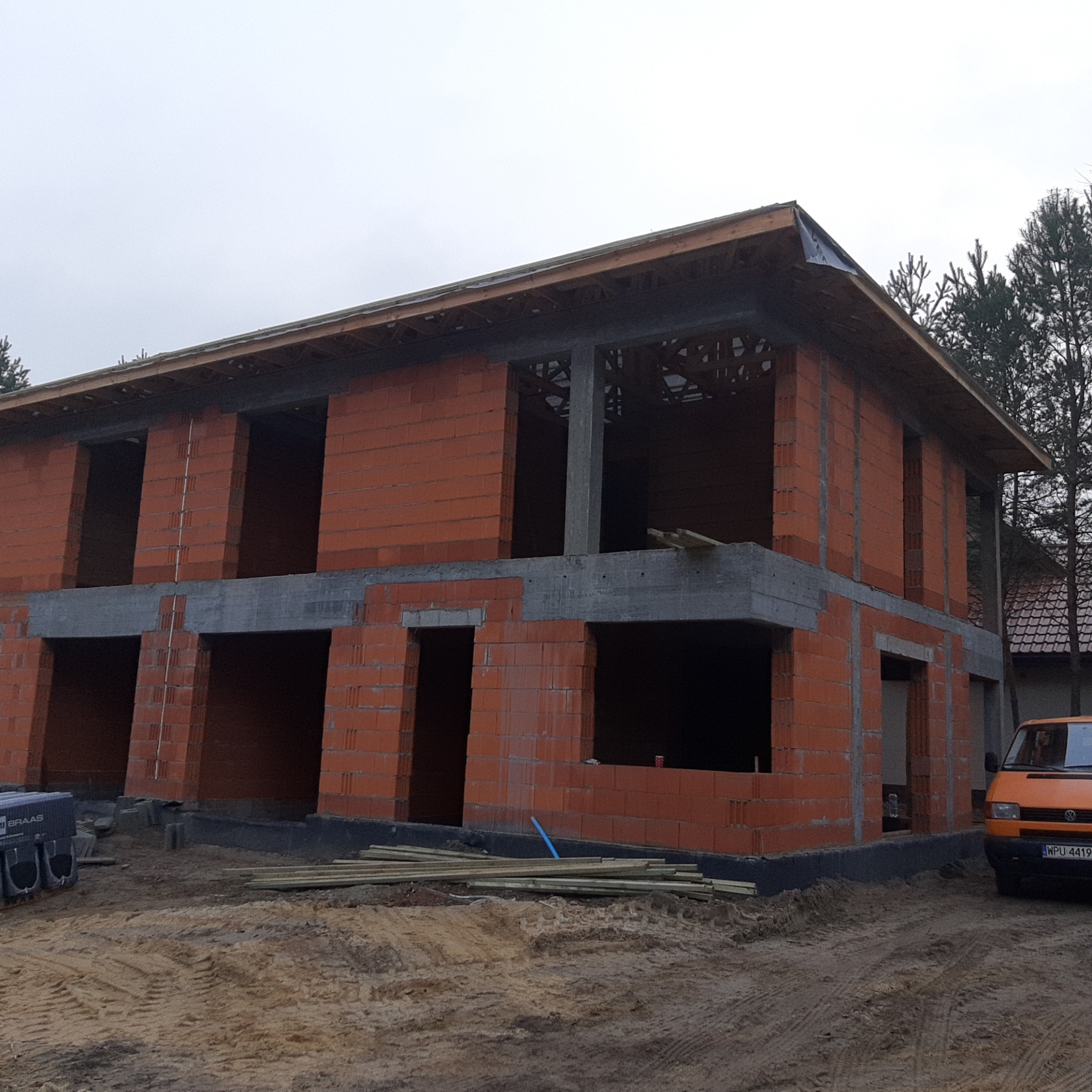 Surowy stan dwukondygnacyjnego domu z czerwonej cegły, widoczne otwory okienne i drzwiowe, częściowo zadaszony, na placu budowy stoi pomarańczowy bus.