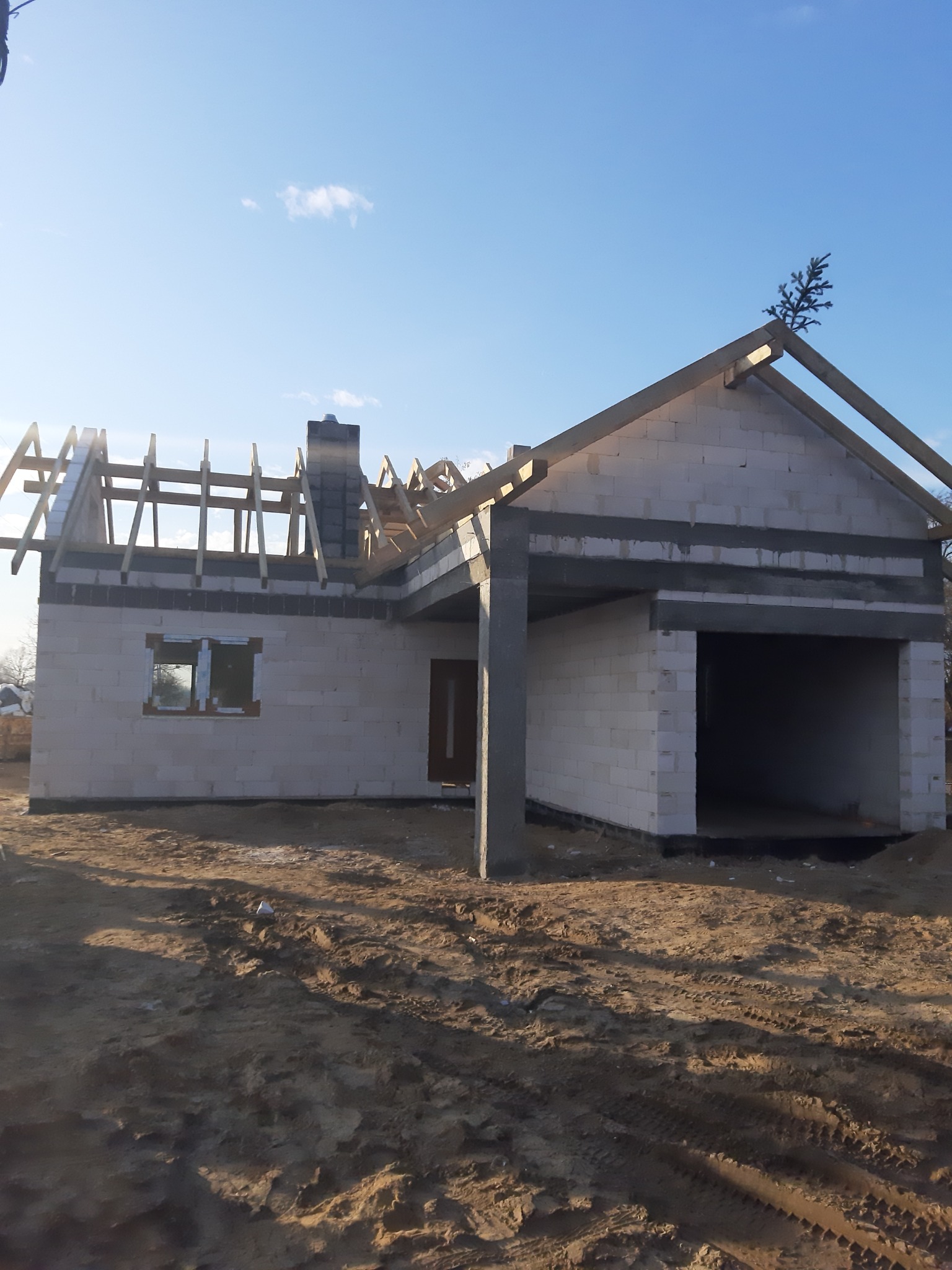 Surowy stan budynku jednorodzinnego z białych bloczków, w trakcie budowy dachu, z małą choinką na szczycie, na tle błękitnego nieba.