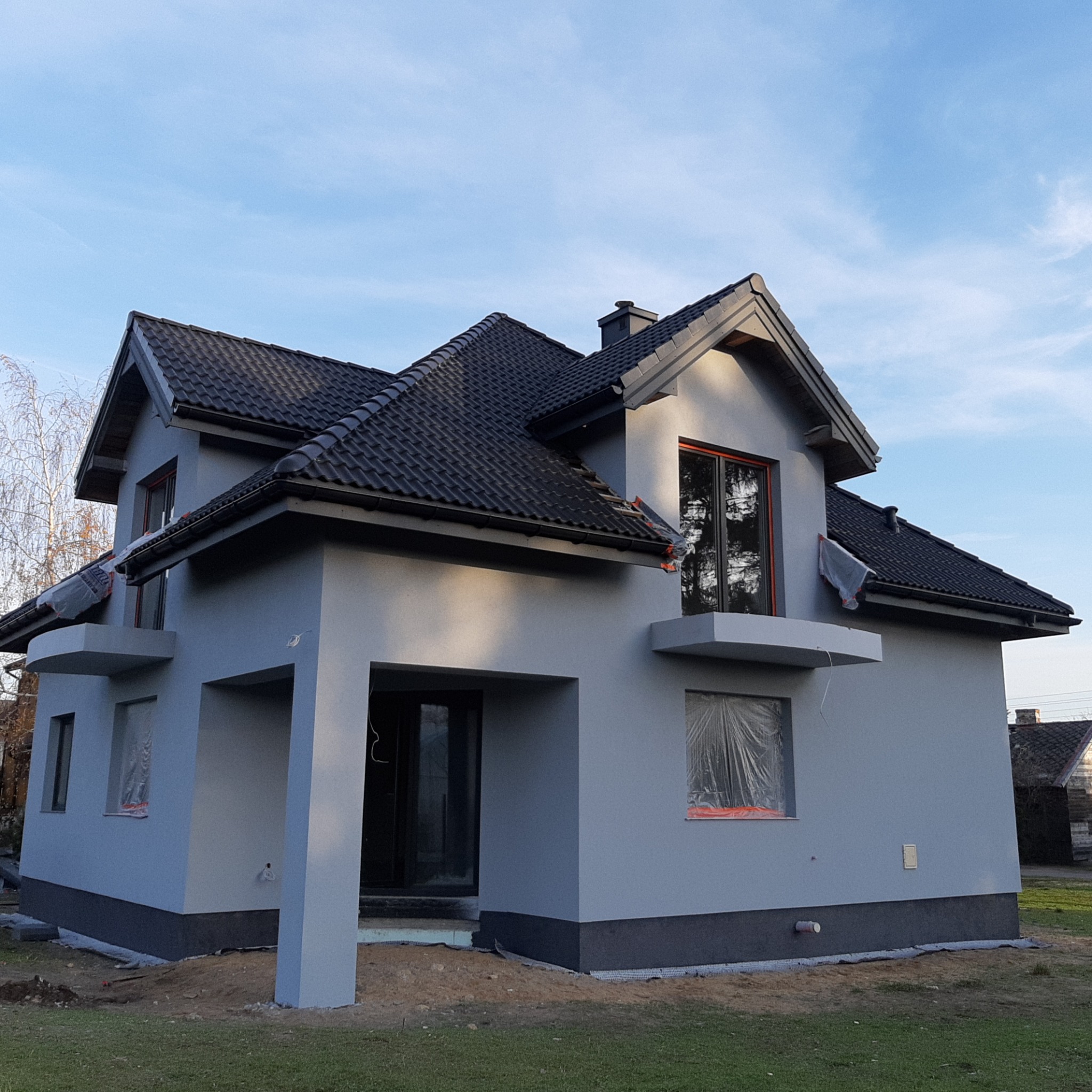 Dom jednorodzinny z szarą elewacją w trakcie prac wykończeniowych, okna zabezpieczone folią, widoczne pozostałości po pracach ziemnych.