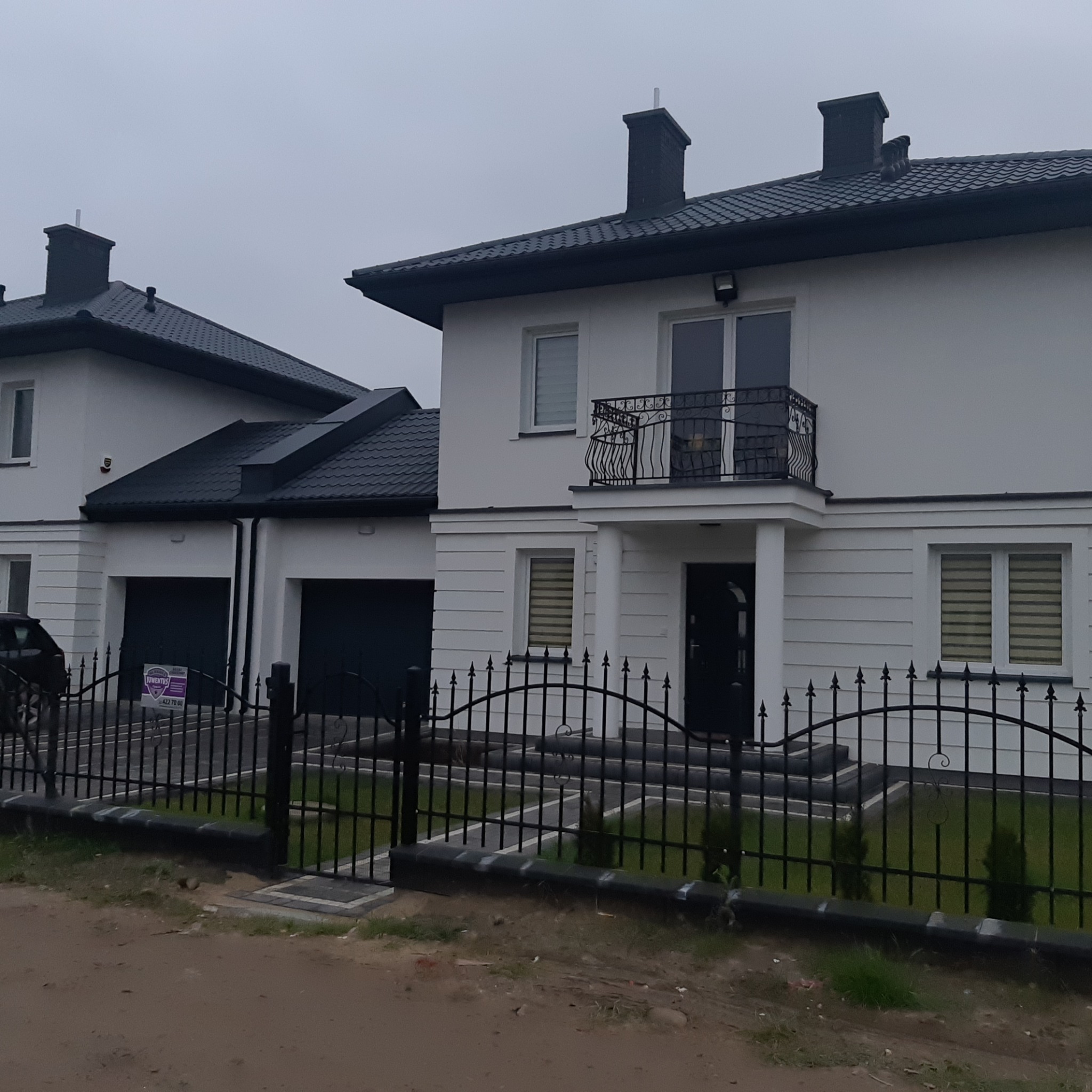 Dwa identyczne, nowoczesne domy z białymi elewacjami, czarnymi dachami i ogrodzeniami, widoczne z zewnątrz posesji w pochmurny dzień.