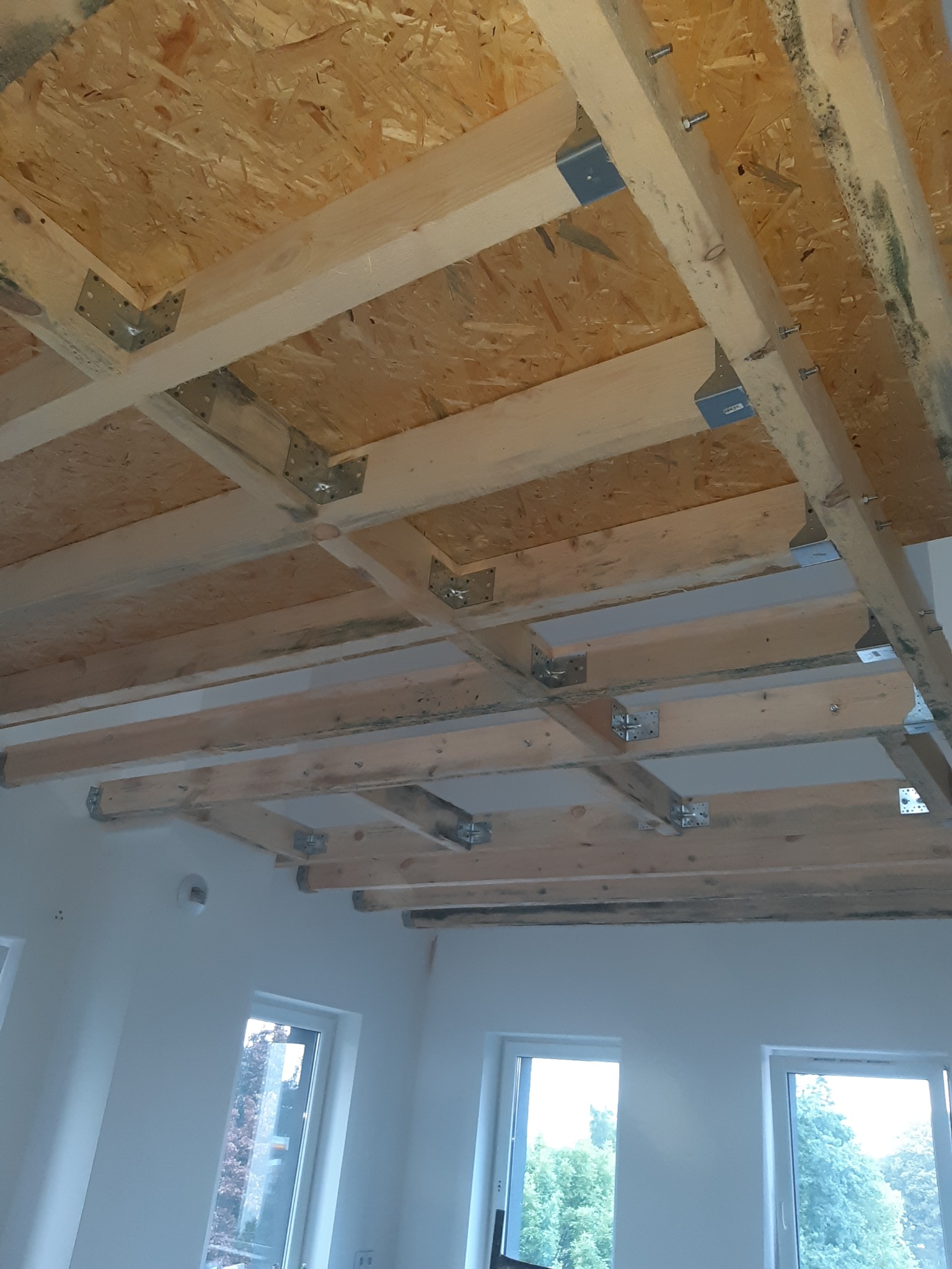 Konstrukcja drewniana poddasza z płyt OSB, mocowana do belek za pomocą metalowych łączników, widok z wnętrza pomieszczenia z oknami.