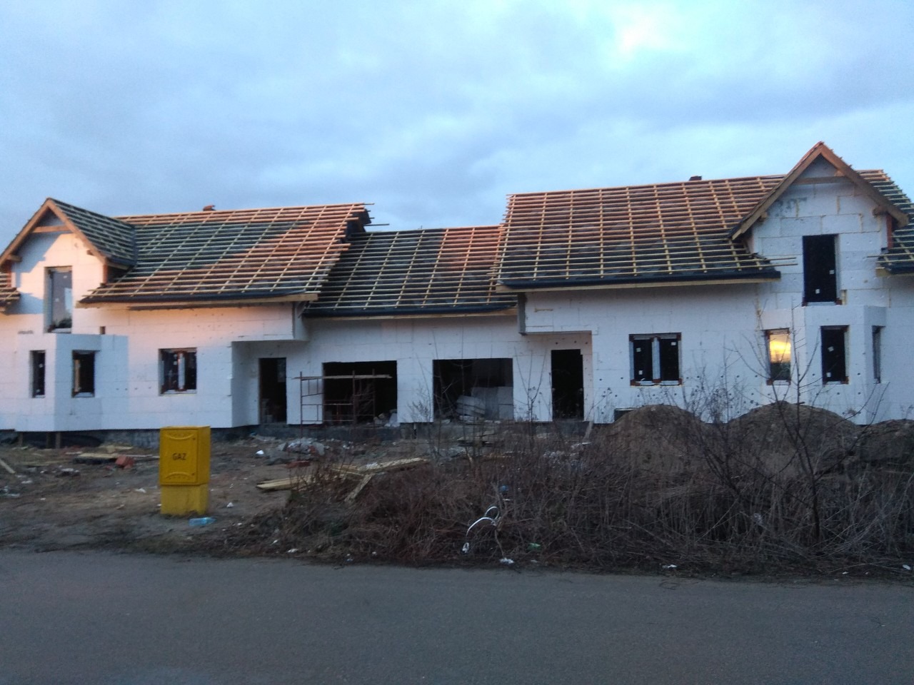 Niedokończony dom jednorodzinny z białego styropianu, widoczne krokwie dachu bez pokrycia, żółta skrzynka gazowa na pierwszym planie.