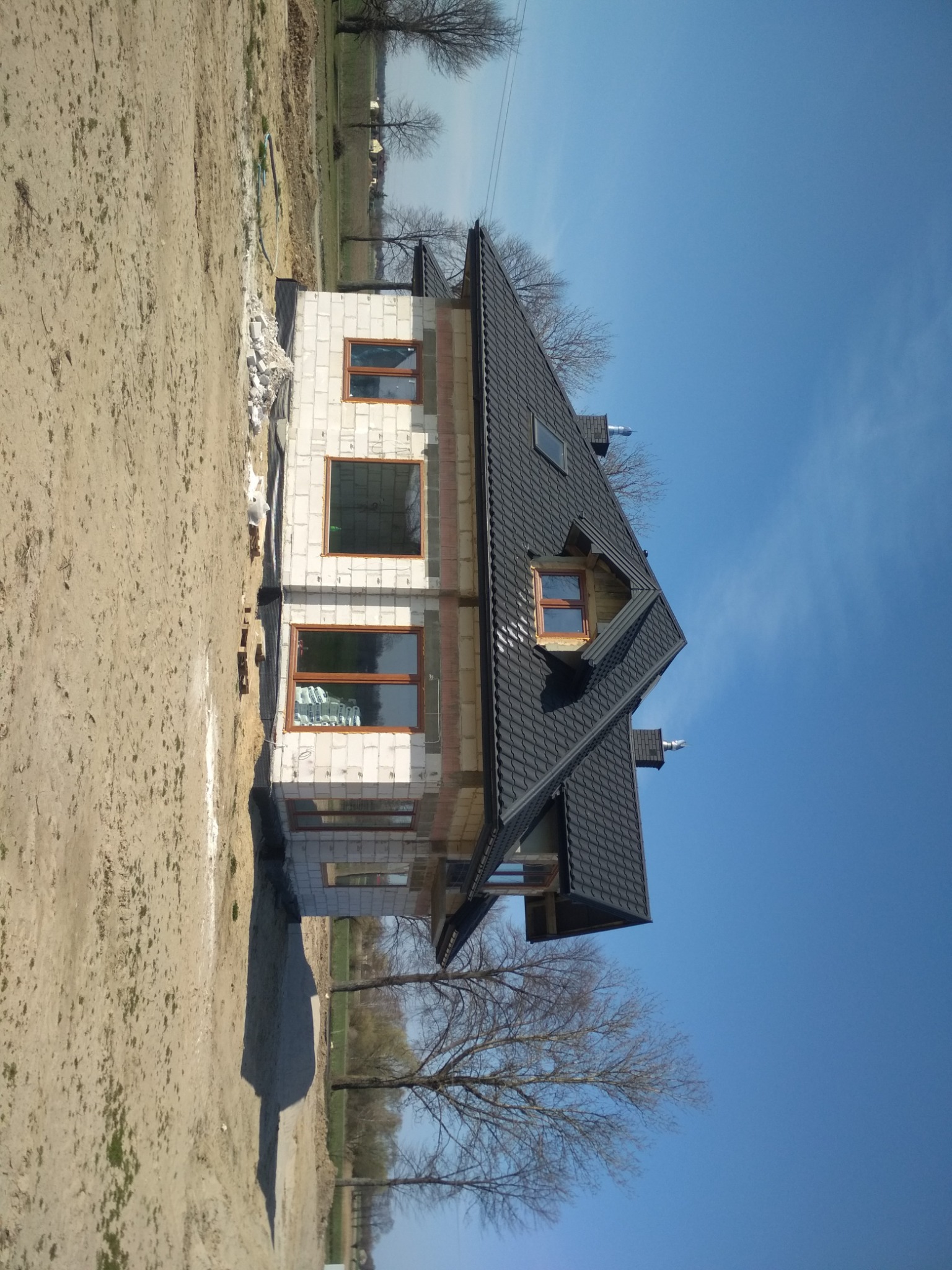 Dwukondygnacyjny dom w stanie surowym otwartym z czarnym dachem, widoczne białe bloczki, okna z brązowymi ramami i niezabezpieczona folią ściana fundamentowa.