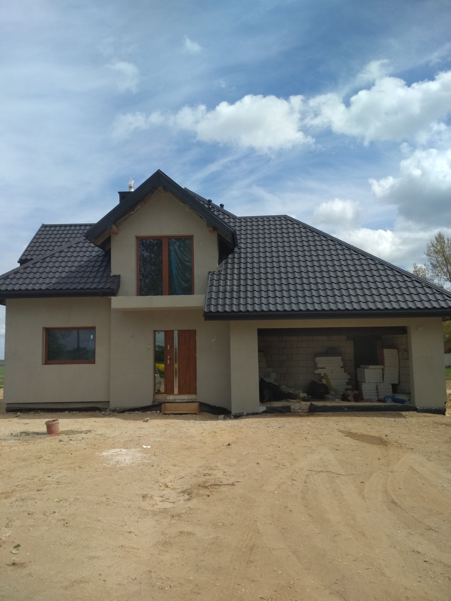 Dom jednorodzinny w trakcie budowy, z szarą elewacją, ciemną dachówką i otwartym garażem wypełnionym bloczkami oraz materiałami izolacyjnymi, na piaszczystym terenie.