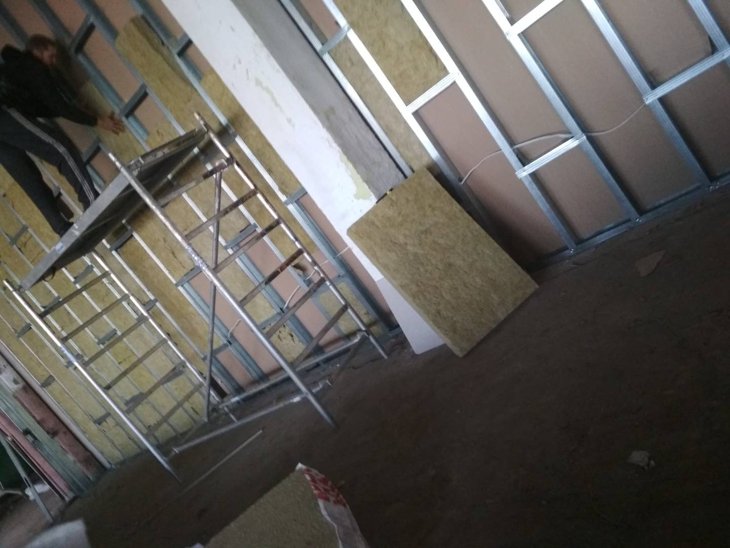 Pracownik na rusztowaniu montuje izolację z wełny mineralnej w metalowym stelażu na ścianie, widoczne płyty kartonowo-gipsowe.