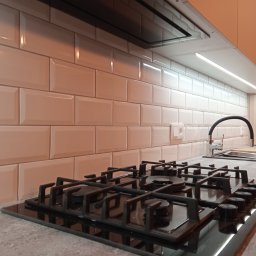 Kolorama Łukasz Mikołajczuk - Białe, błyszczące płytki ułożone w cegiełkę na ścianie nad blatem kuchennym z czarną płytą gazową i zlewozmywakiem z czarną baterią.