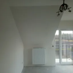 Pokój na poddaszu po wyrównaniu ścian, z zamontowanym grzejnikiem i lampą sufitową, widoczne ślady niedoskonałości na ścianach, okno balkonowe z widokiem na zewnątrz.