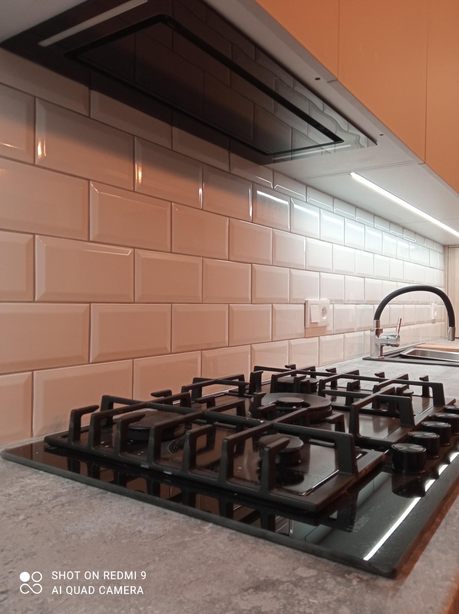 Białe, błyszczące płytki ułożone w cegiełkę na ścianie nad blatem kuchennym z czarną płytą gazową i zlewozmywakiem z czarną baterią.