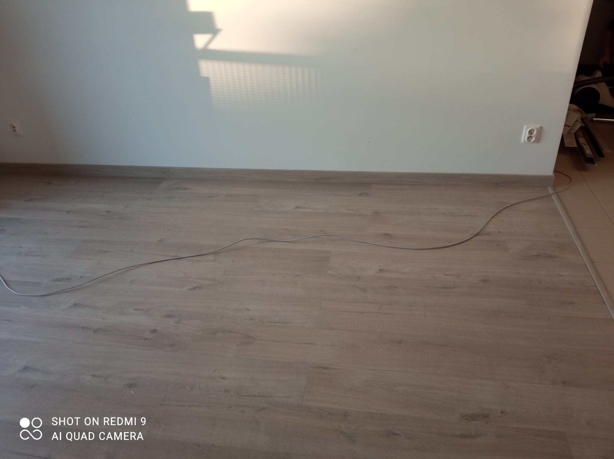 Jasne panele podłogowe ułożone w pokoju, widoczny kabel elektryczny na podłodze i cień na ścianie.
