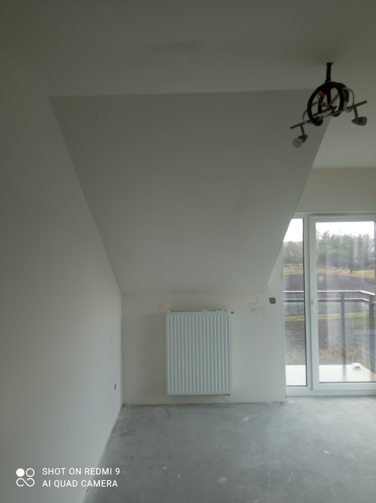 Pokój na poddaszu po wyrównaniu ścian, z zamontowanym grzejnikiem i lampą sufitową, widoczne ślady niedoskonałości na ścianach, okno balkonowe z widokiem na zewnątrz.