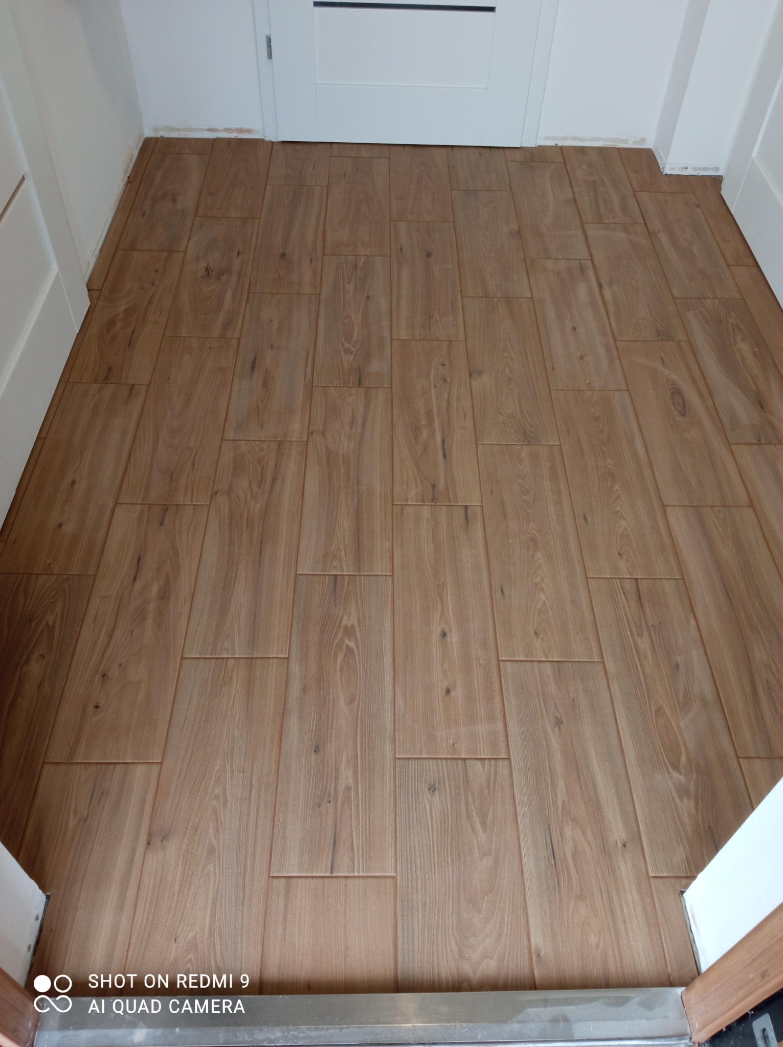 Ułożone panele podłogowe imitujące drewno w wąskim korytarzu, widoczne białe futryny drzwiowe i listwa progowa.