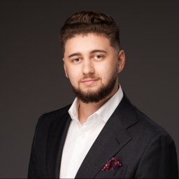 MS LEGIT Kancelaria Adwokacka Michał Słota - Młody mężczyzna z zadbaną brodą i ułożonymi włosami, ubrany w ciemny garnitur, białą koszulę i czerwoną poszetkę w biało-niebieskie wzory, pozuje na szarym tle.