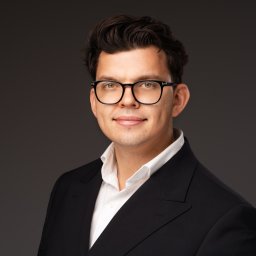 MS LEGIT Kancelaria Adwokacka Michał Słota - Profesjonalny portret uśmiechniętego mężczyzny w okularach i czarnym garniturze, z ciemnymi, ułożonymi włosami, na gładkim, ciemnoszarym tle.