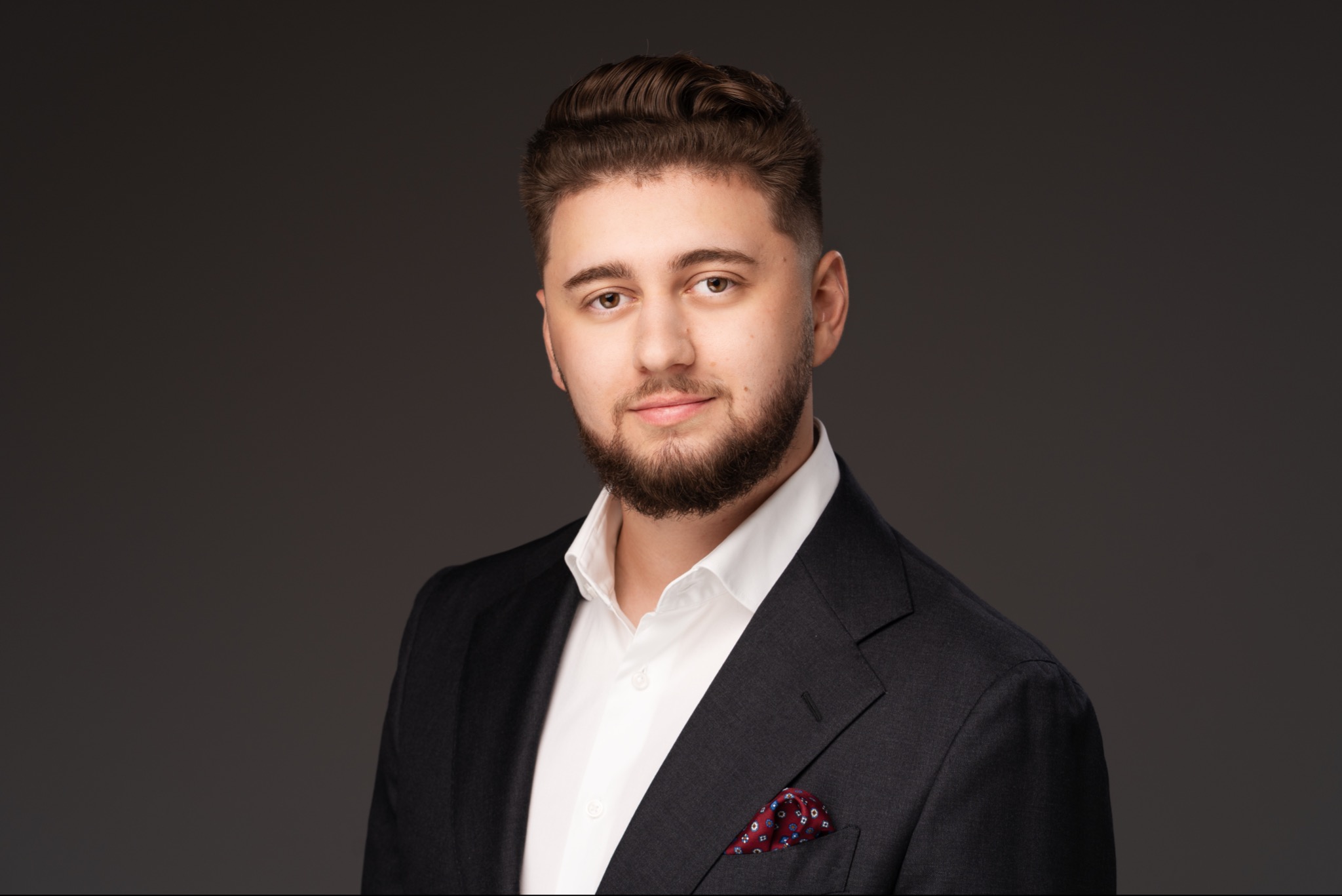 Młody mężczyzna z zadbaną brodą i ułożonymi włosami, ubrany w ciemny garnitur, białą koszulę i czerwoną poszetkę w biało-niebieskie wzory, pozuje na szarym tle.