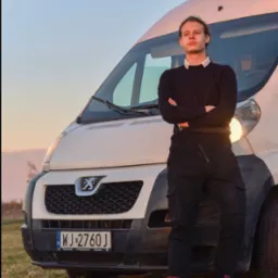 Młody mężczyzna oparty o przód białego busa Peugeot z polską tablicą rejestracyjną WJ 2760J, stojącego na trawie o zmierzchu.