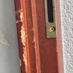 MERANTI KRZYSZTOF CZEKAJŁO - Uszkodzony, brązowy profil drewniany drzwi z widocznym zamkiem w kolorze starego złota i śladami odpryskującej farby oraz ubytkami drewna.