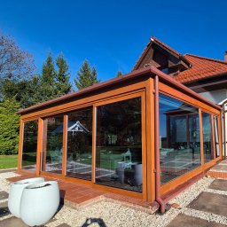 MERANTI KRZYSZTOF CZEKAJŁO - Zadaszony taras z drewnianą konstrukcją i dużymi oknami, widok z zewnątrz na tle domu z czerwoną dachówką i zieleni. Dwa białe donice na pierwszym planie.