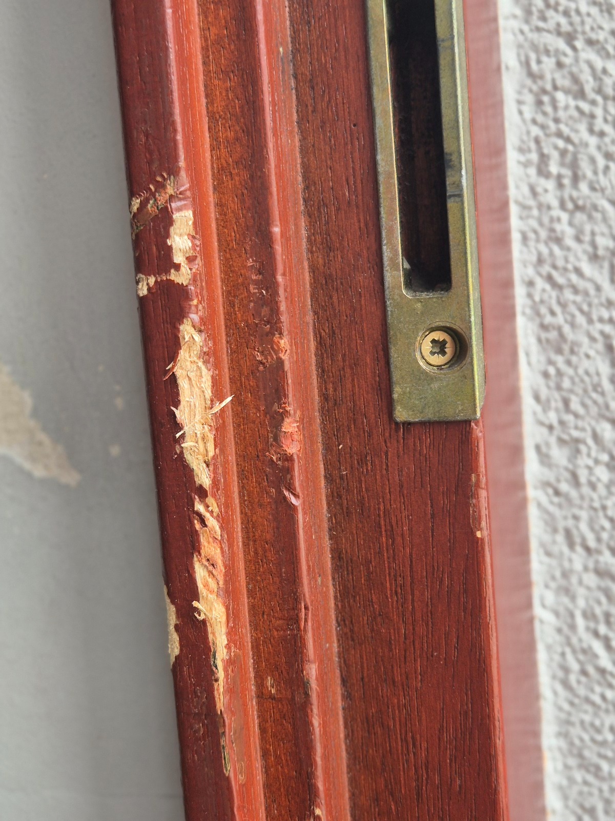 Uszkodzony, brązowy profil drewniany drzwi z widocznym zamkiem w kolorze starego złota i śladami odpryskującej farby oraz ubytkami drewna.