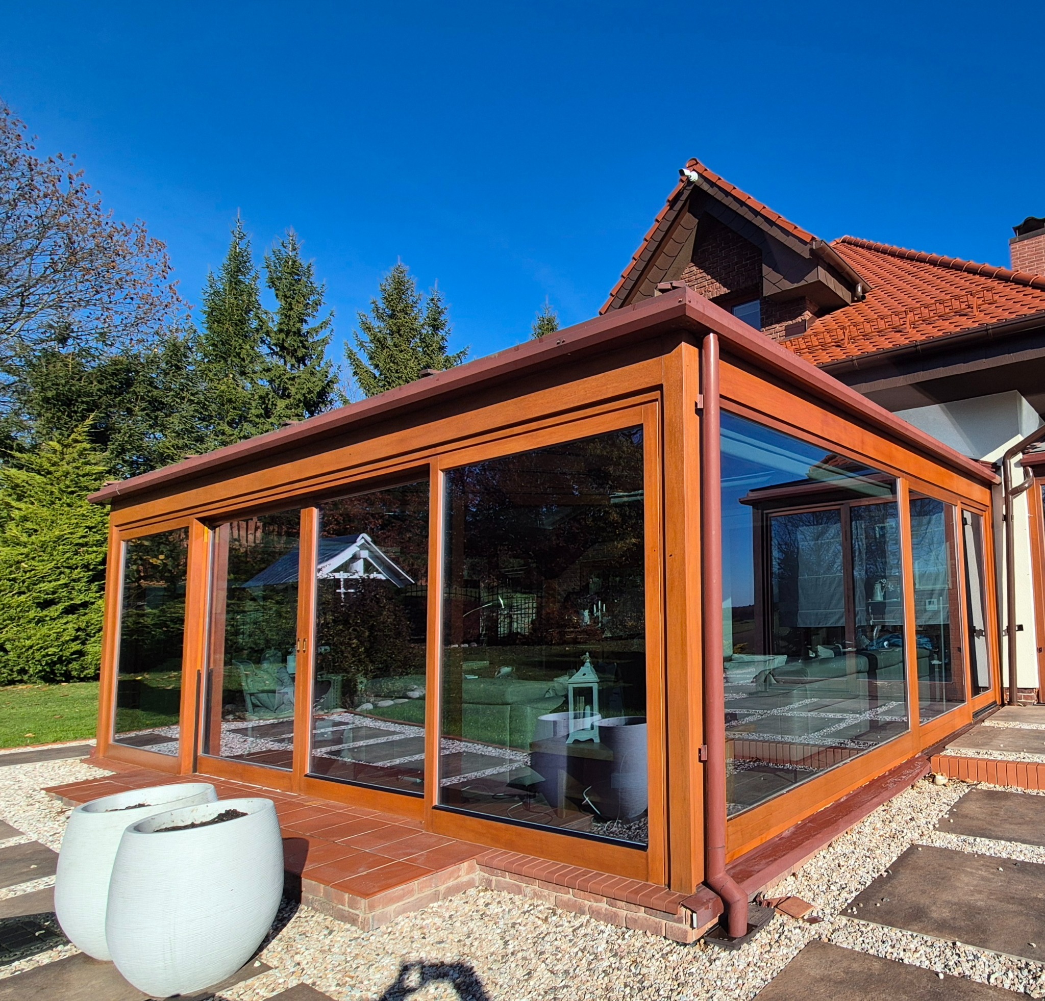 Zadaszony taras z drewnianą konstrukcją i dużymi oknami, widok z zewnątrz na tle domu z czerwoną dachówką i zieleni. Dwa białe donice na pierwszym planie.