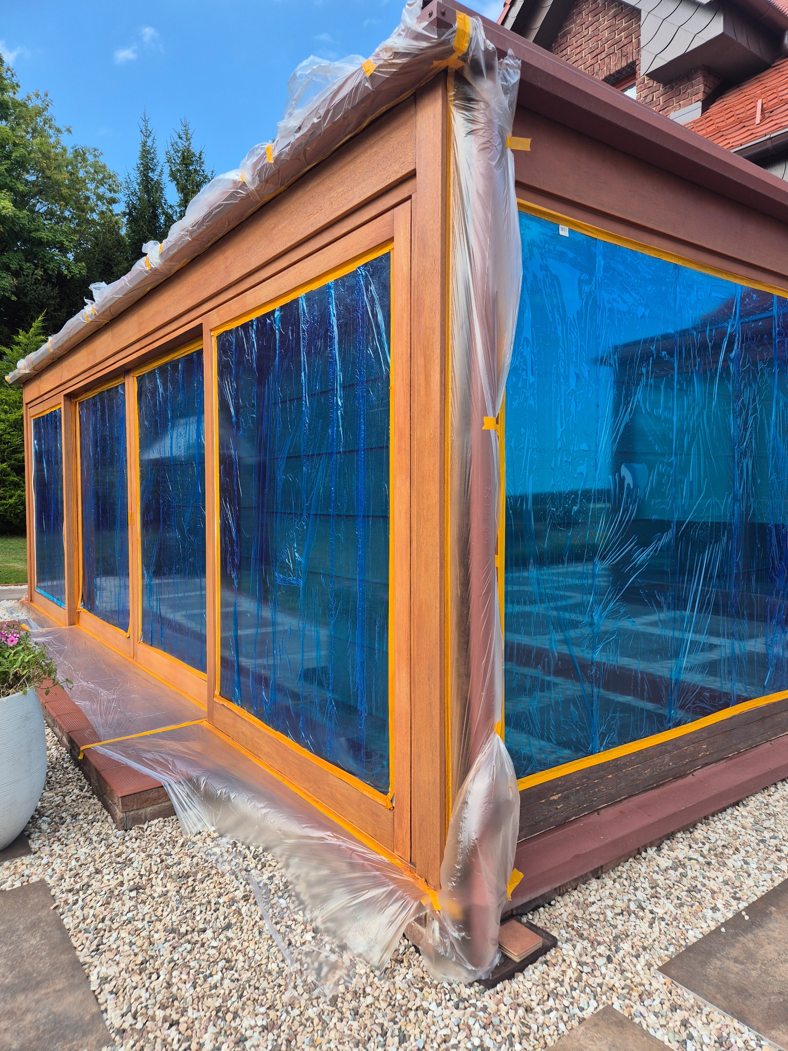 Przygotowanie do malowania drewnianej konstrukcji z oknami, zabezpieczonej folią i taśmą malarską we Wrocławiu. Widok z zewnątrz, w tle zieleń.