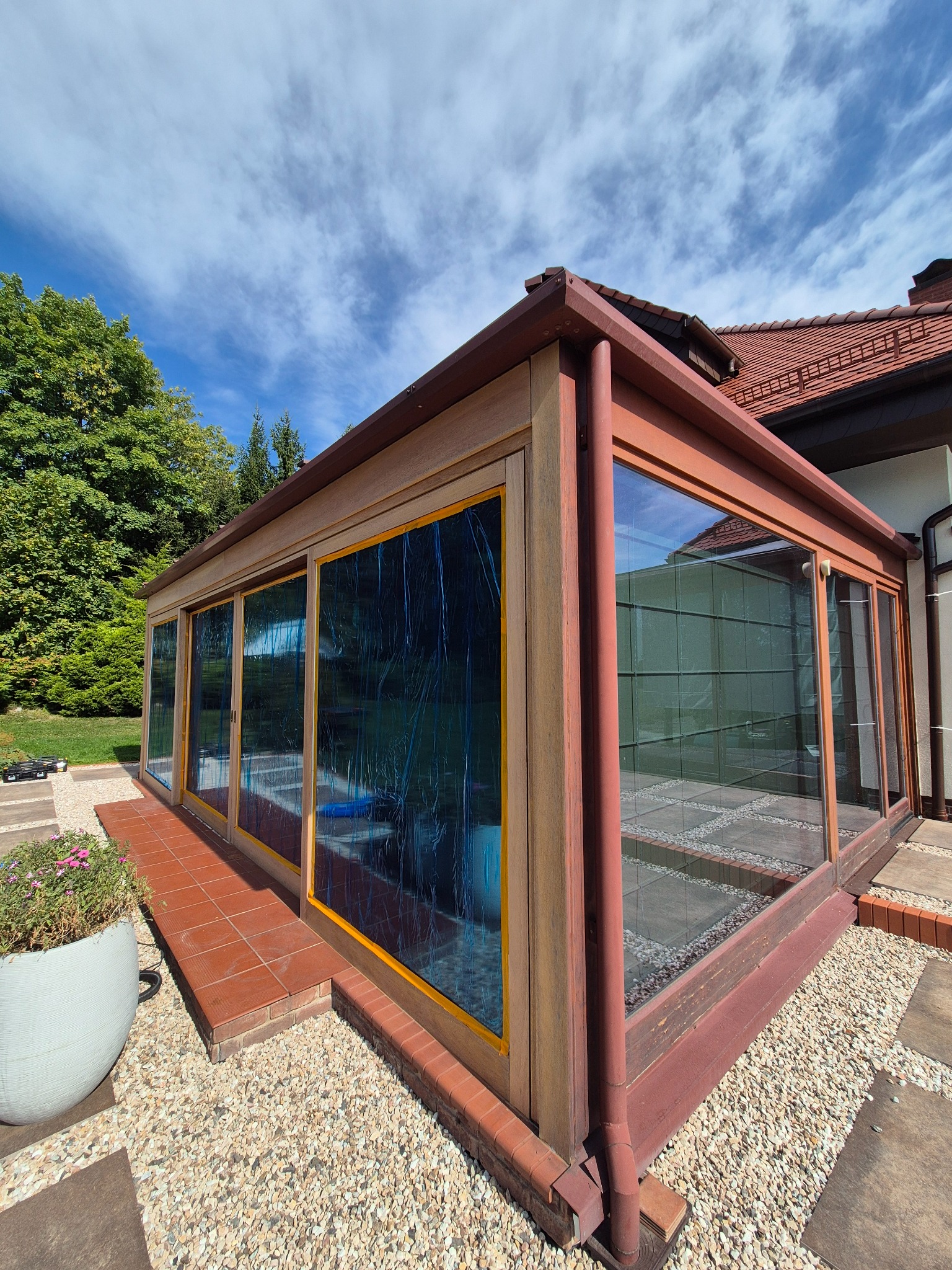 Zadaszenie tarasu z drewnianą konstrukcją i dużymi przeszkleniami, widok z zewnątrz na tle ogrodu i błękitnego nieba z chmurami. Element architektury ogrodowej.