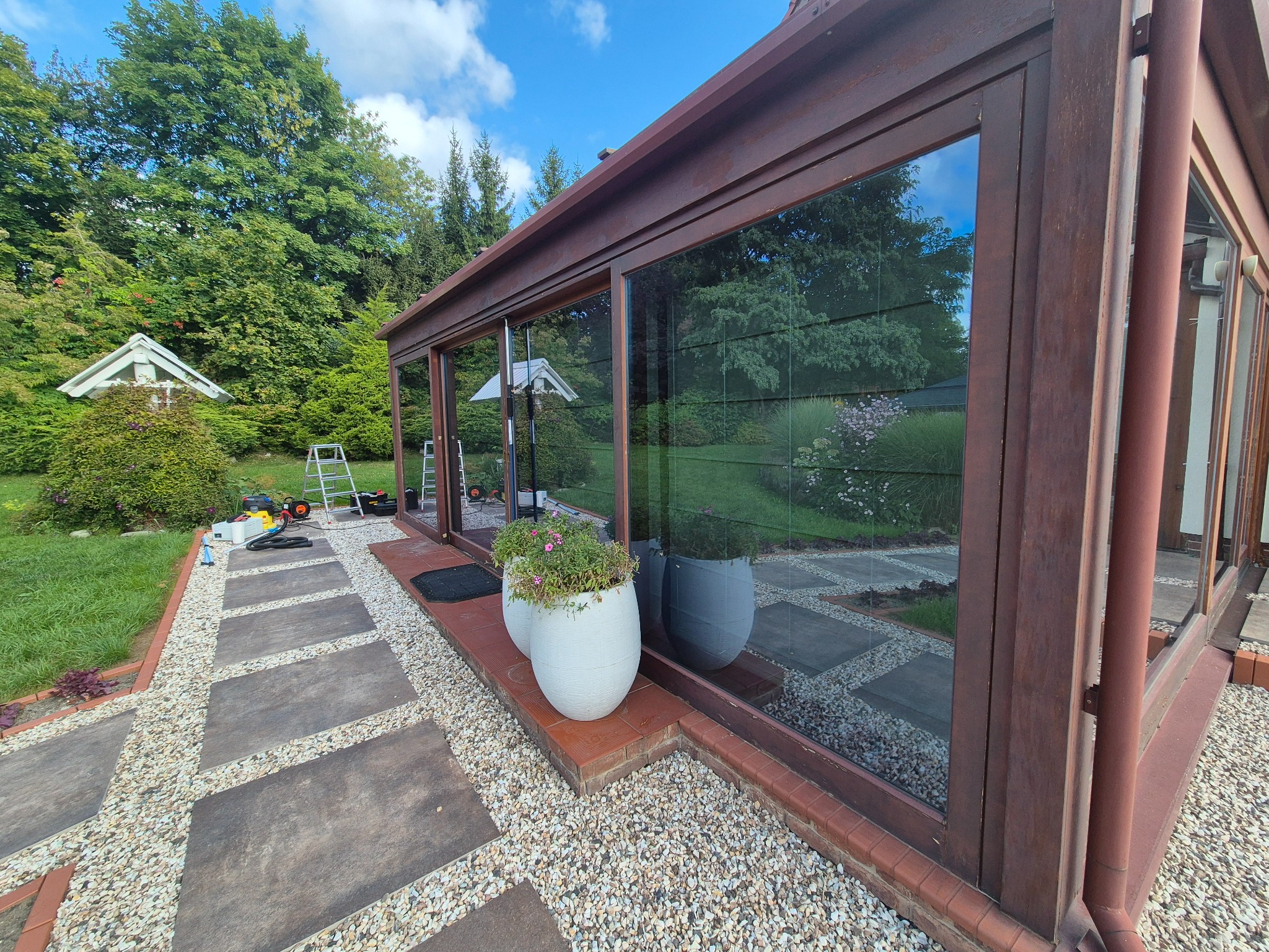 Rustykalna, przeszklona weranda z brązowymi ramami okiennymi, otoczona zielenią ogrodu. Na pierwszym planie donice z kwiatami i ścieżka z kamiennych płyt.