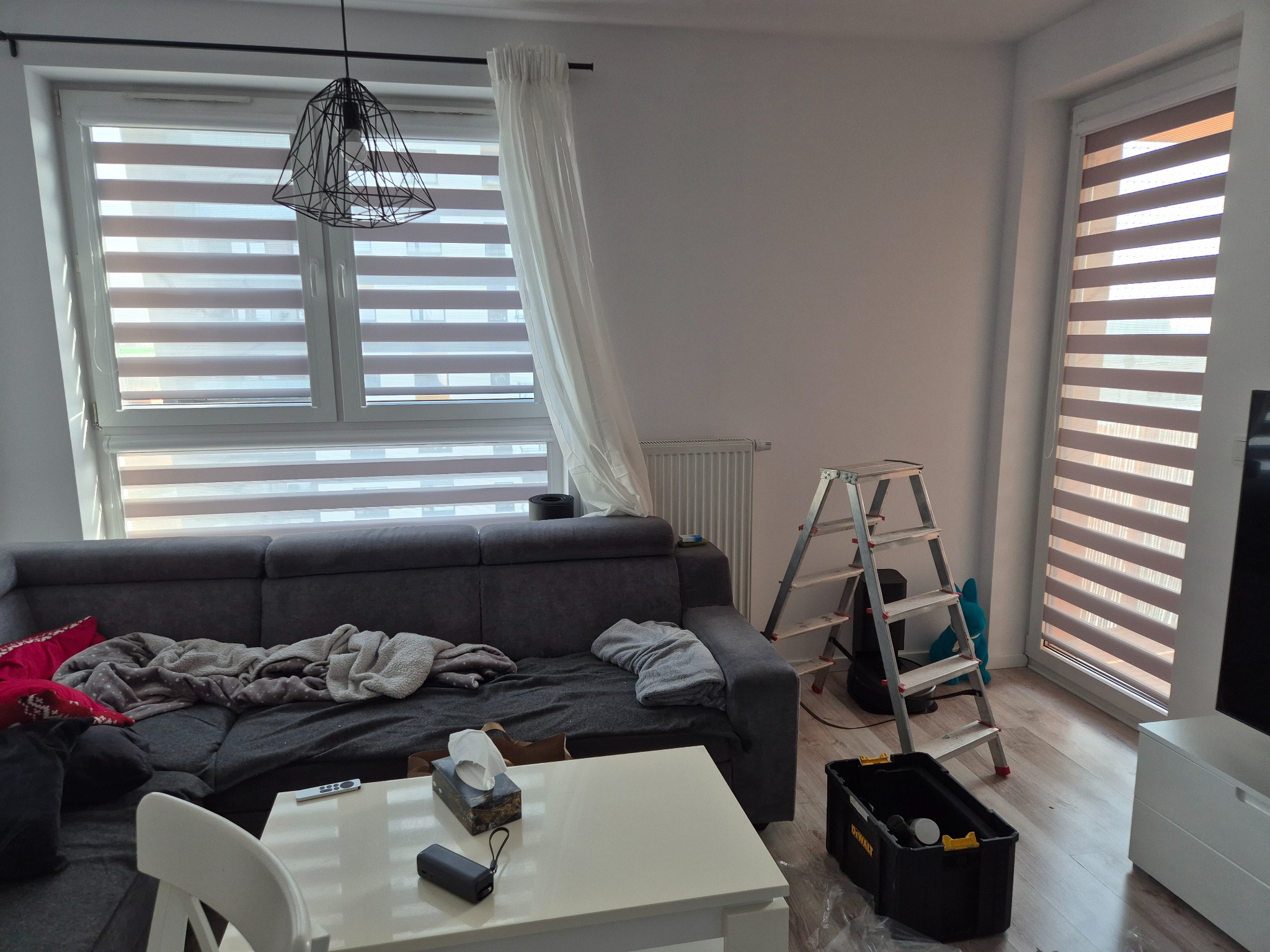 Pokój z oknami przesłoniętymi roletami dzień-noc, drabina i skrzynka narzędzi sugerują prace remontowe. Lampa wisząca i sofa dodają wnętrzu charakteru.