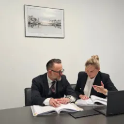 Dwoje prawników w biurze analizuje dokumenty przy biurku, na którym leży laptop. Na ścianie wisi obraz z motywem Dublina.