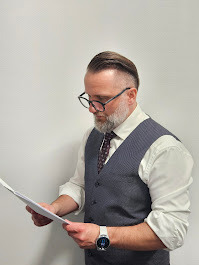 Mężczyzna w okularach i kamizelce analizuje dokumenty, na nadgarstku widoczny smartwatch, na jednolitym tle. Ujęcie od pasa w górę.